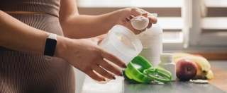 Athletic woman in sportswear with measuring spoon in her hand puts portion of whey protein powder into a shaker on wooden table with amino acid white capsules, bananas and apple, making protein drink.