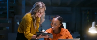 Two businesswomen at a table working together while using a laptop and tablet.