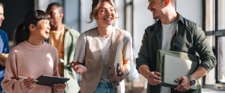 A group of young professionals walk together in a bright, modern office space, smiling and chatting casually while holding notebooks and tablets.