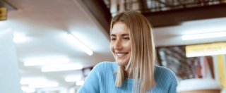 Education, university and happy woman in library with laptop, research and books for school project or exam. Smile, internet and college phd student studying with technology and elearning on campus.