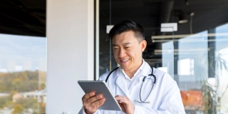 Cheerful and friendly Asian in a modern clinic, online video call, uses a tablet, to communicate with patients.